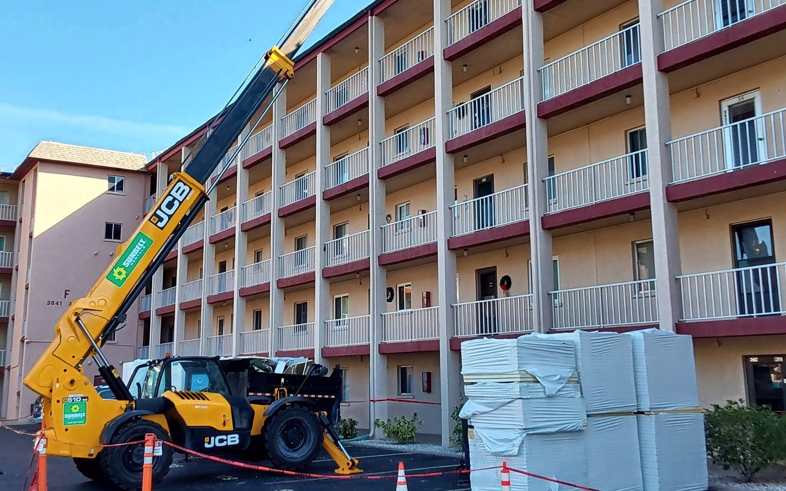Telehandler lifting roofing materials from a taped-off work zone beside a commercial building